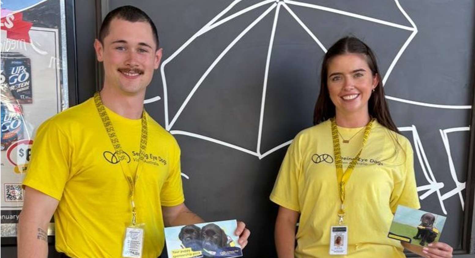 A young man and a young woman in yellow Seeing Eye Sogs t-shirts, holding up Seeing Eye Dog postcards