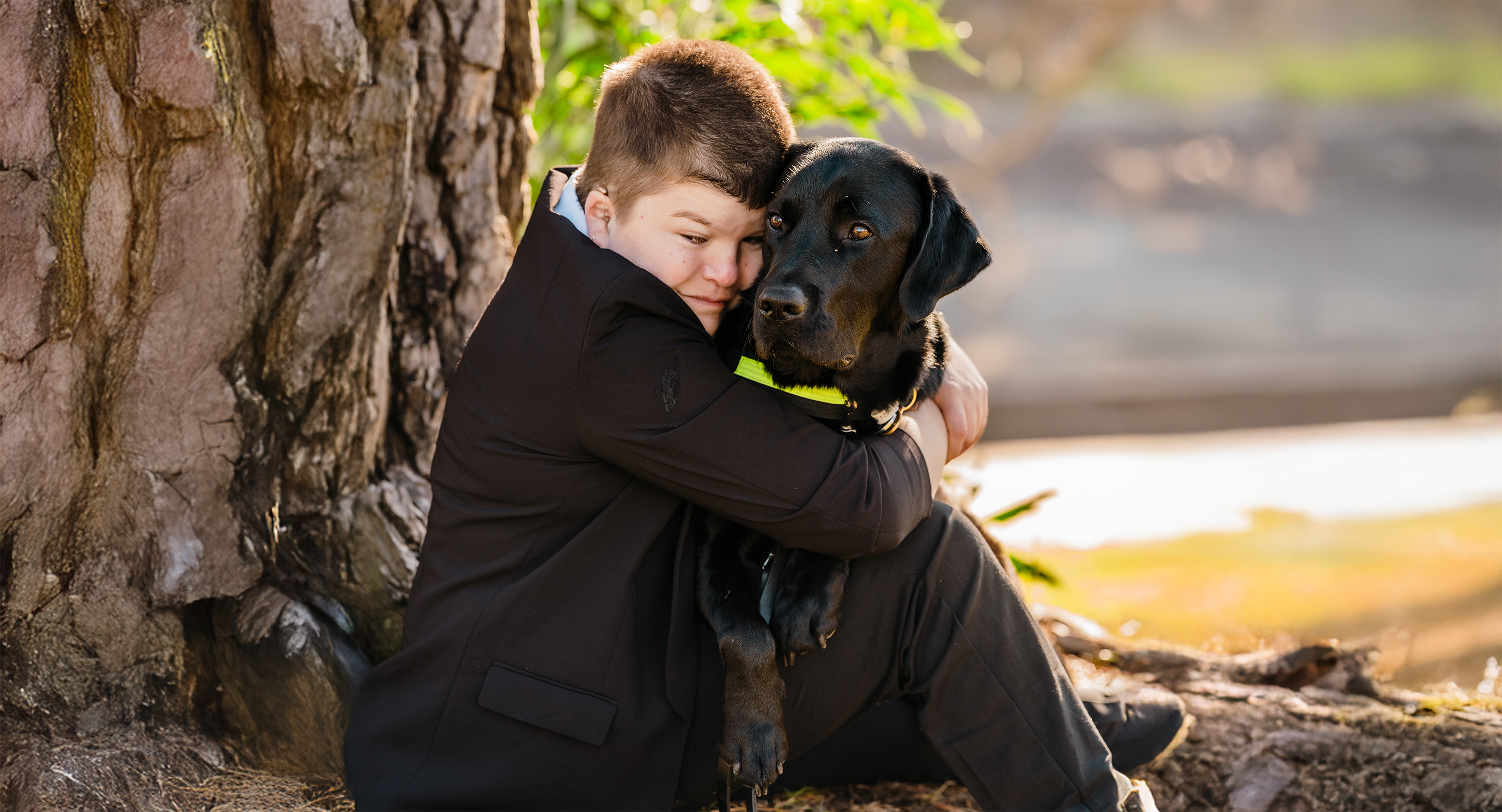 Alayna Campbell sits against a tree and cuddles her Seeing Eye Dog, Joey, who is a black Labrador. Behind them is some green foliage and both her and Joey look towards the camera.