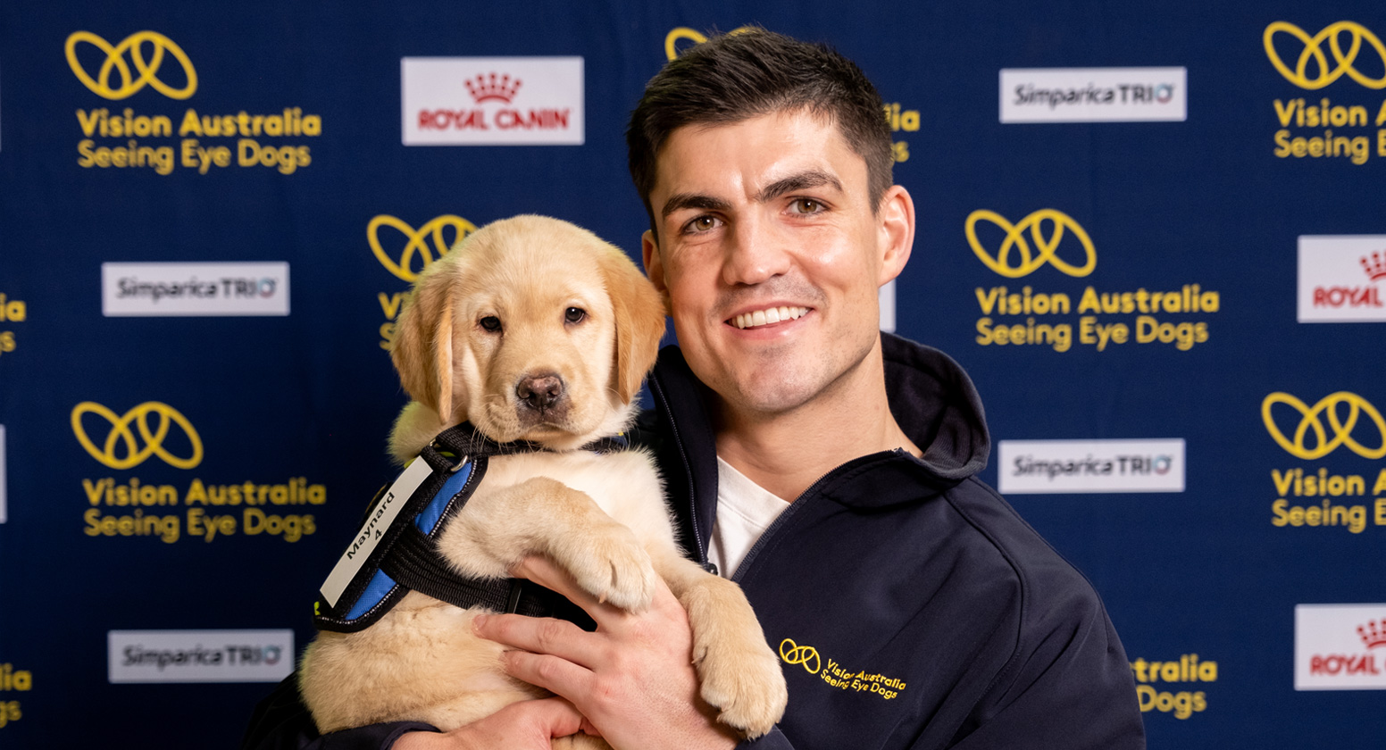 Seeing Eye Dogs ambassador and AFL Premiership player Brayden Maynard holds a yellow Seeing Eye Dogs puppy, which is wearing its training coat.