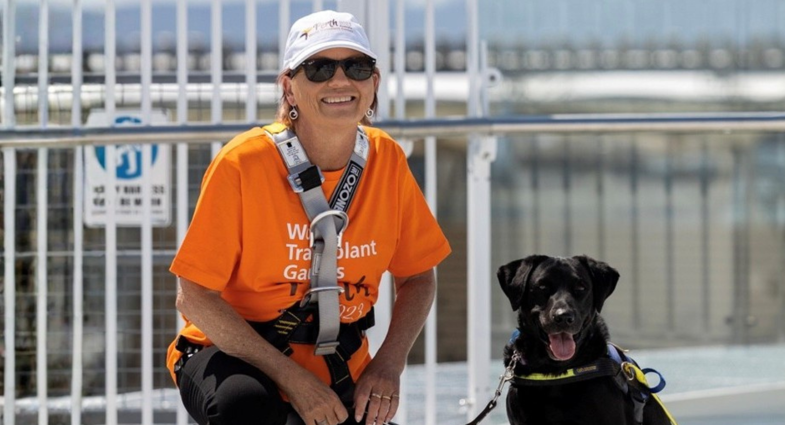 Seeing Eye Dogs client, Colleen, sits next to her black Labrador Seeing Eye Dog, Inca, while smiling at the camera. She is wearing an orange t-shirt. 