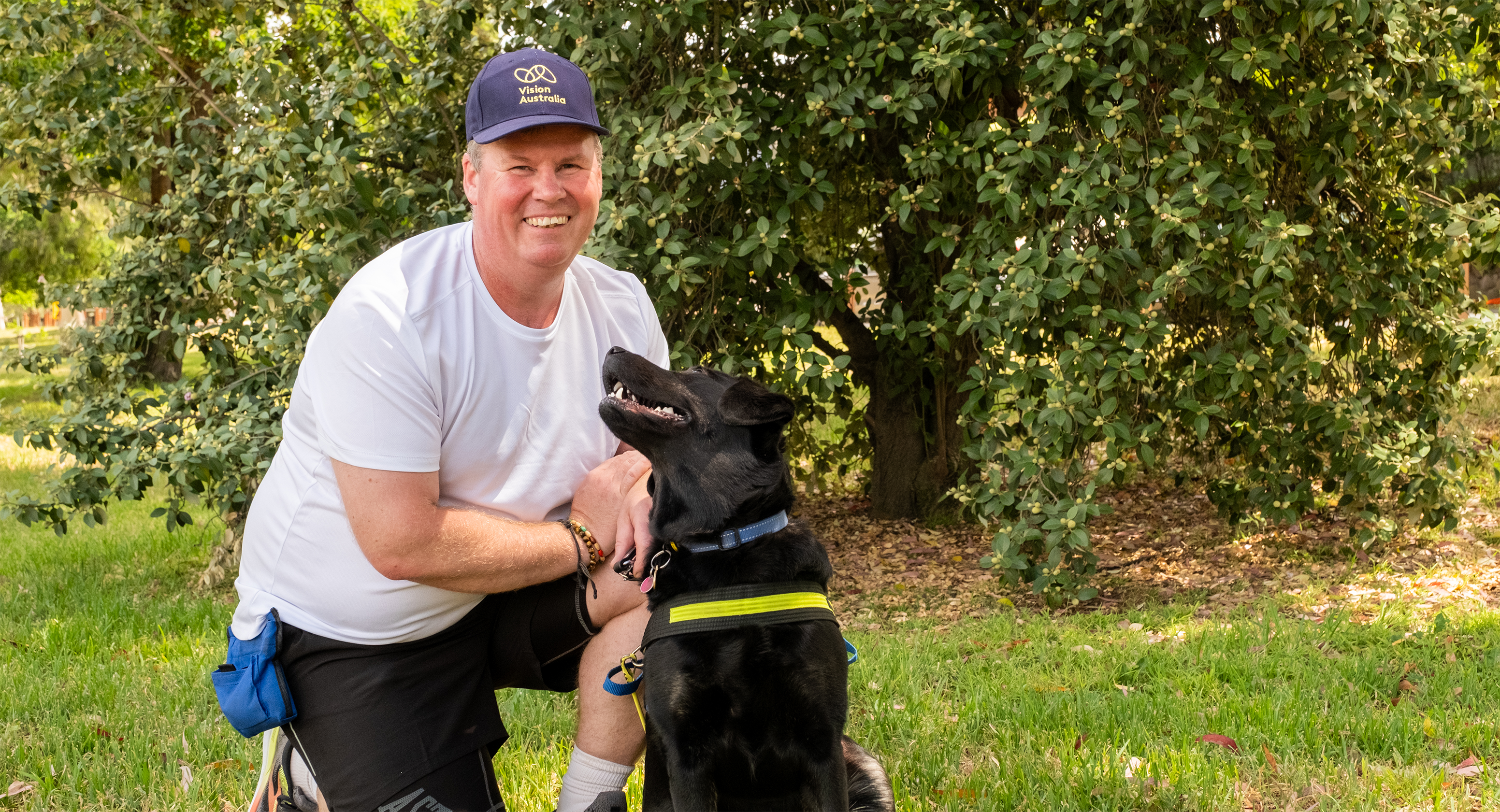 Dale Pierce is pictured kneeling on the grass in a white t-shirt, black running shorts and a Vision Australia hat, smiling at the camera. Next to him is his Seeing Eye Dog Yana, a black Labrador, who has her head turned up looking towards him. Behind them is some green foliage.