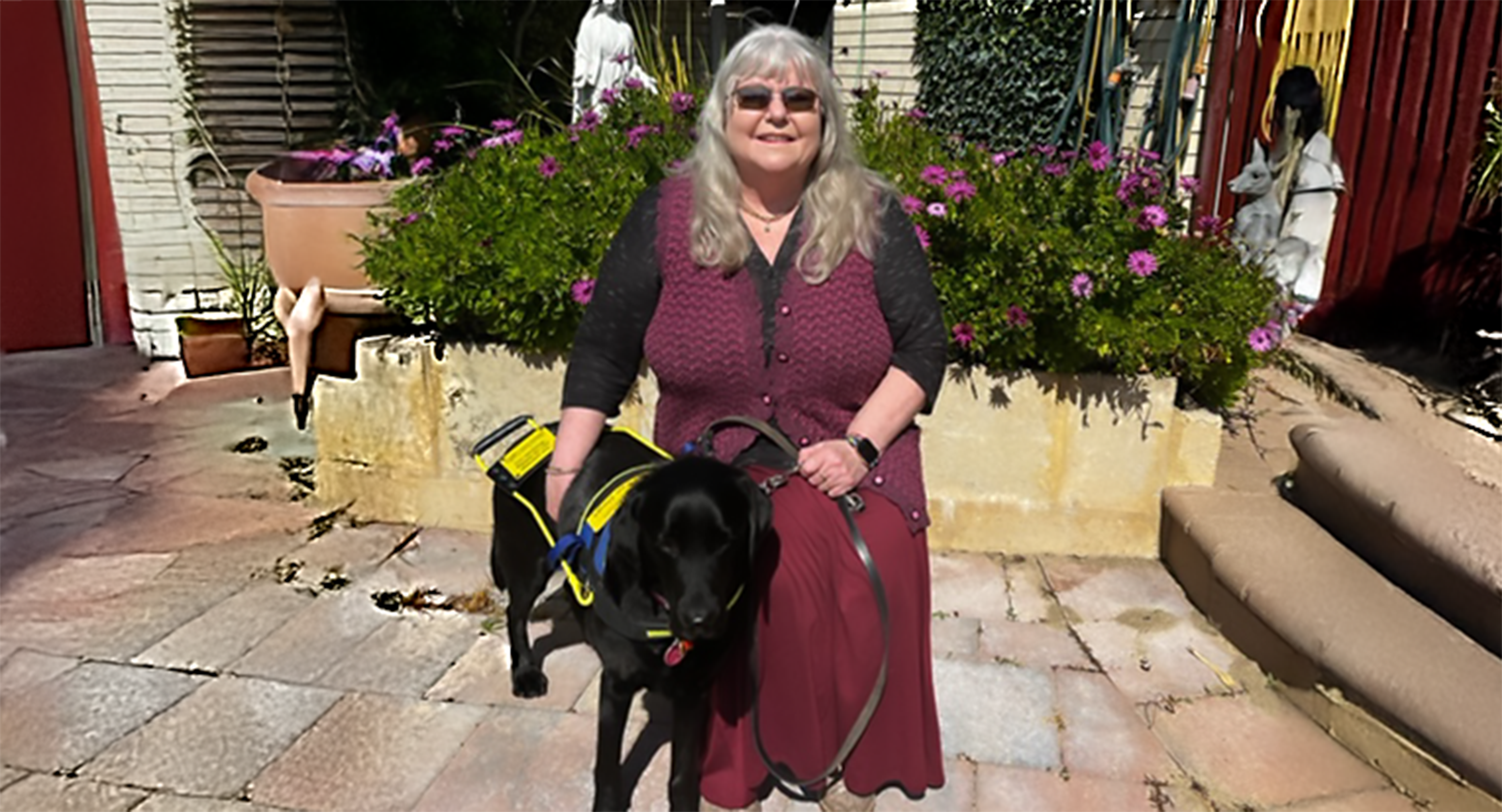 Geraldine Lane is pictured sitting in the sun, wearing a maroon coloured dress while patting her Seeing Eye Dog, Noodle. Noodle is a black Labrador and is looking towards the camera. Behind them appears to be a garden, including a flower bed with purple flowers and a garden statue.