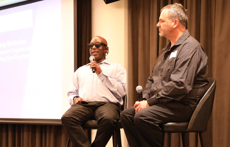 Two men are on a stage, one of the men is holding a microphone and wearing dark glassed