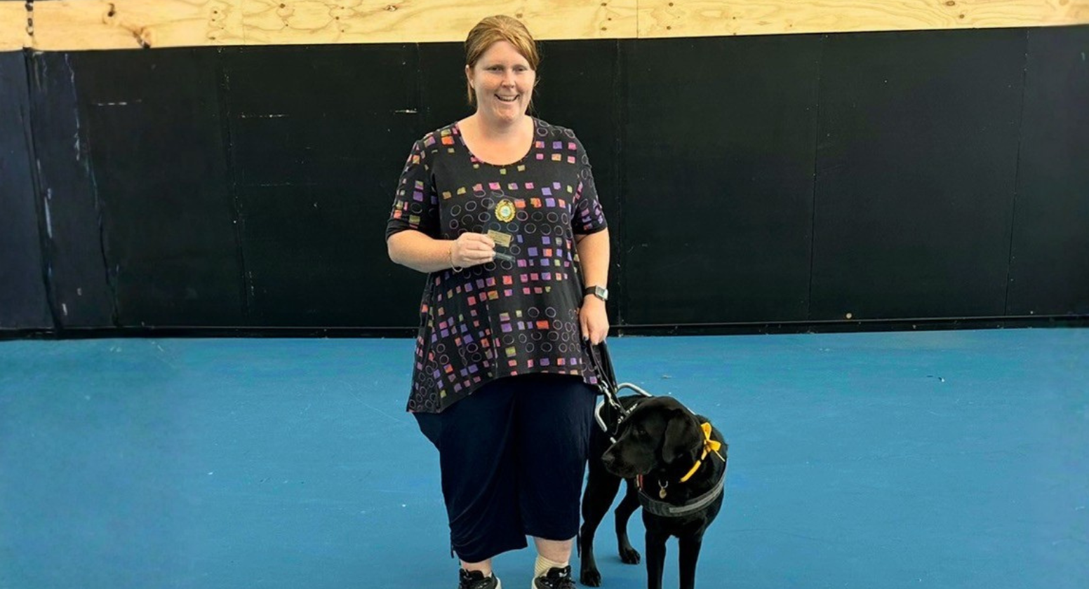 eeing Eye Dogs client, Isabella, stands next to her black Labrador Seeing Eye Dog, Ira, while smiling at the camera. She is standing on a tennis court while holding an award.