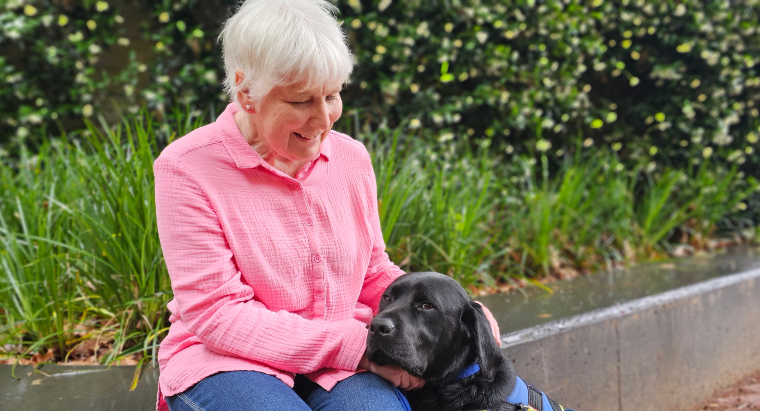 Seeing Eye Dogs client, Kate, sits next to her black Labrador Seeing Eye Dog, Kramer. She sits in front of some greenery in a pink shirt and smiles down at Kramer. 
