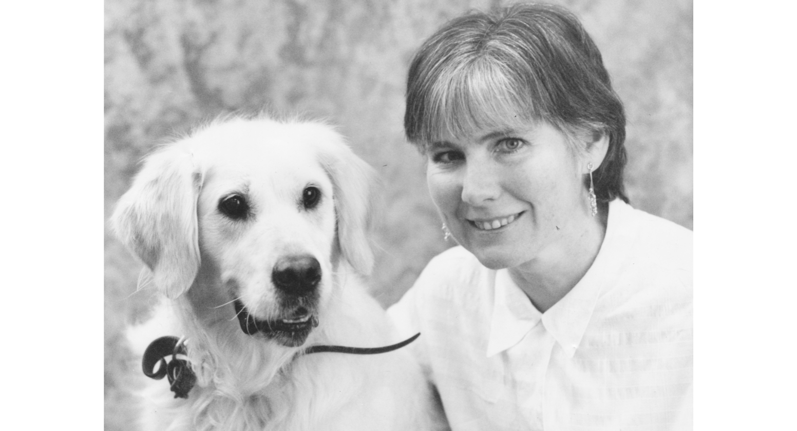 A black and white image of Seeing Eye Dogs client, Kate, next to one of her previous Golden Retriever Seeing Eye Dogs, Le Tisse, and smiles at the camera. 