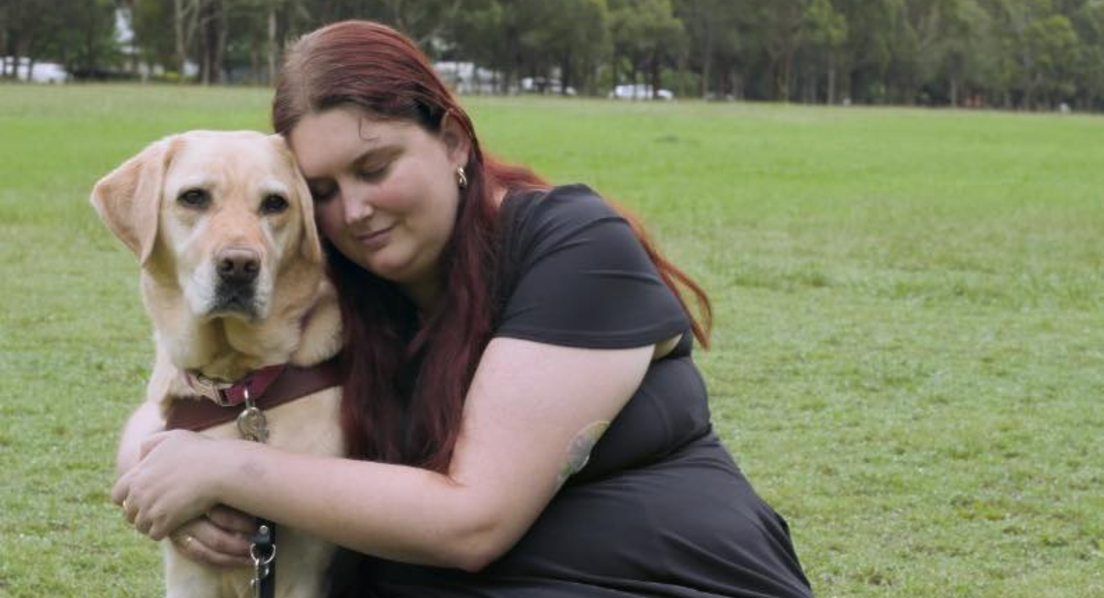 Seeing Eye Dogs client, Kirsten, cuddles her yellow Labrador Seeing Eye Dog, Kimba, on a green lawn. 