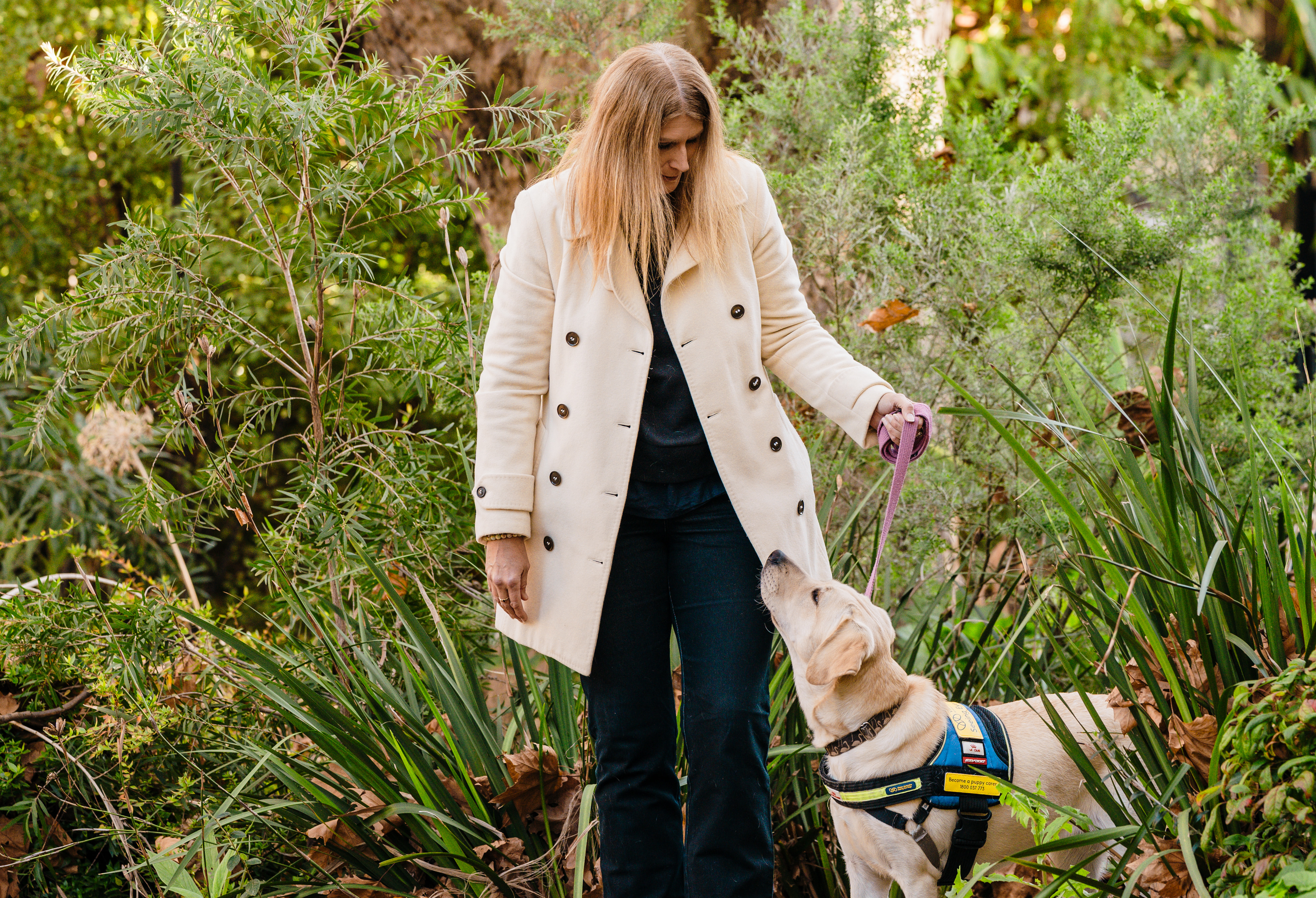 Kylie is in a park holding the leash of a yellow Seeing Eye Dogs puppy wearing its training coat, who is looking up at her. 