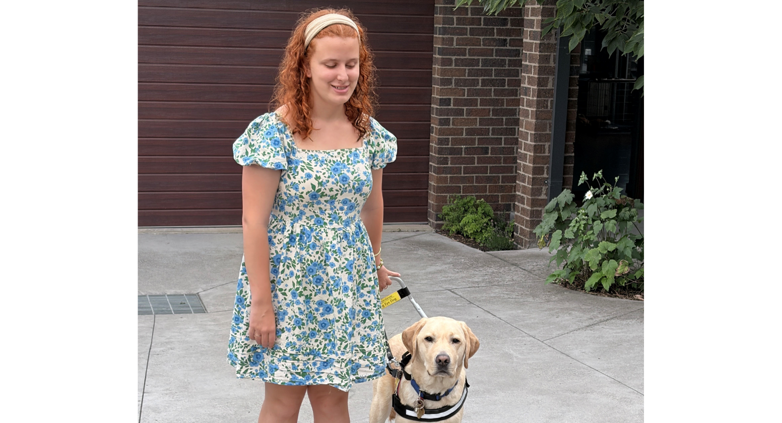 Seeing Eye Dogs client, Lilly, stands next to her yellow Labrador Seeing Eye Dog, Teddy. She wears a blue floral dress and smiles towards the camera. 