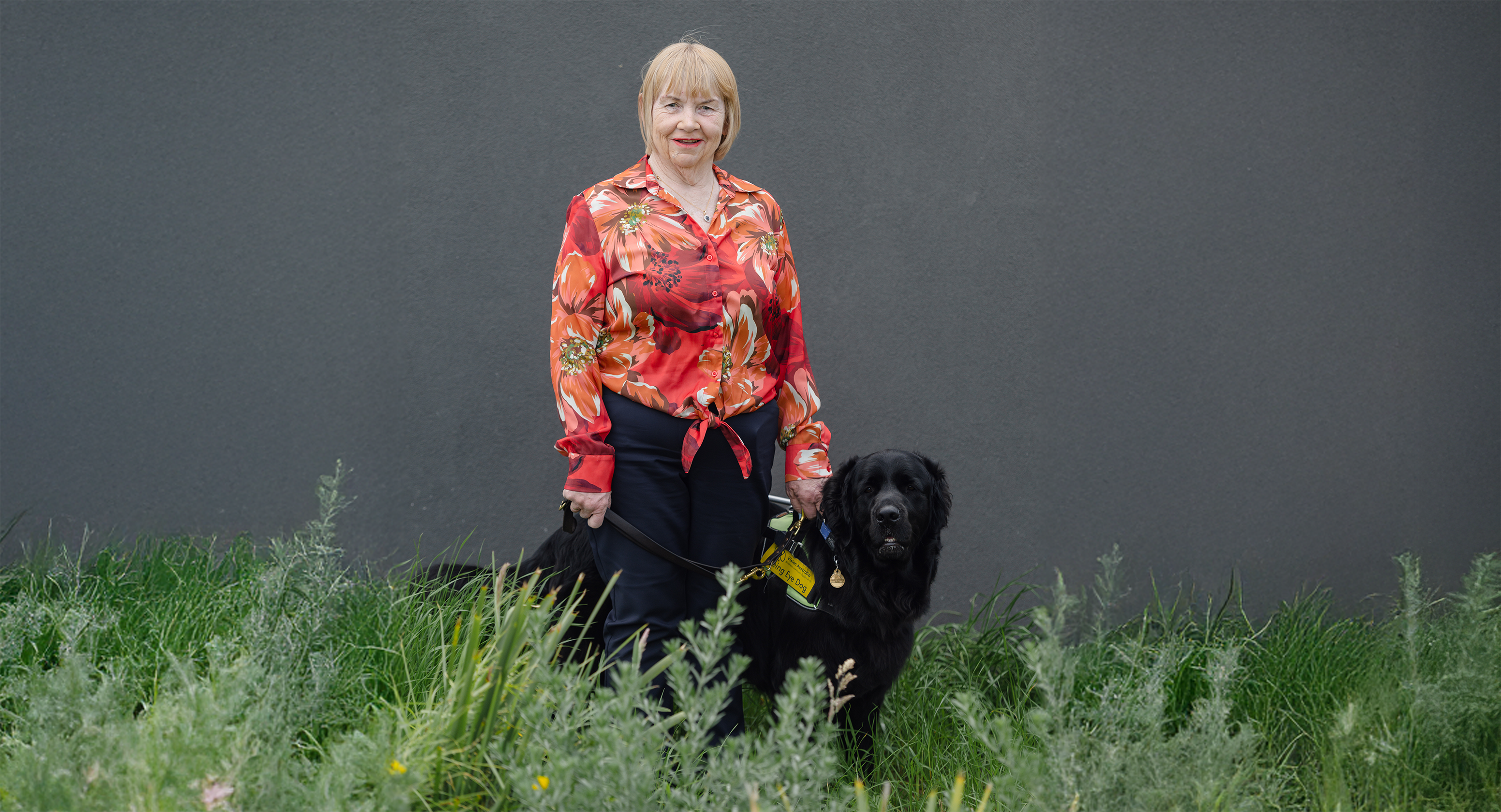 Linda Blaik is pictured standing in some long grass with her Seeing Eye Dog, Kevy. Linda wears a bright red floral shirt that contrasts well against the dark grey wall behind her. Kevy is a mix of a black Labrador and Golden Retriever, and they both look towards the camera.