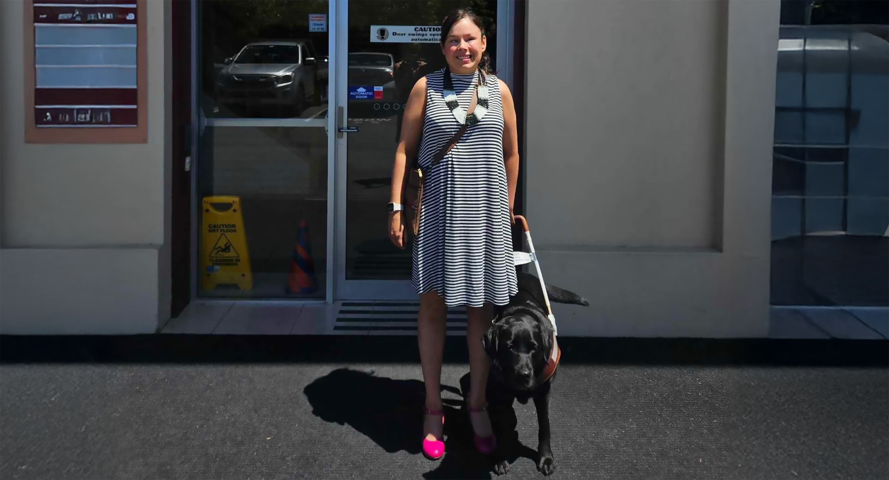 Lizzie Eastham stands in front of a Vision Australia Radio building and smiles towards the camera. She is pictured in a black and white striped dress and bright pink heels, and has her Seeing Eye Dog, Lacey, next to her. Lacey is a black Labrador and is also facing the camera.