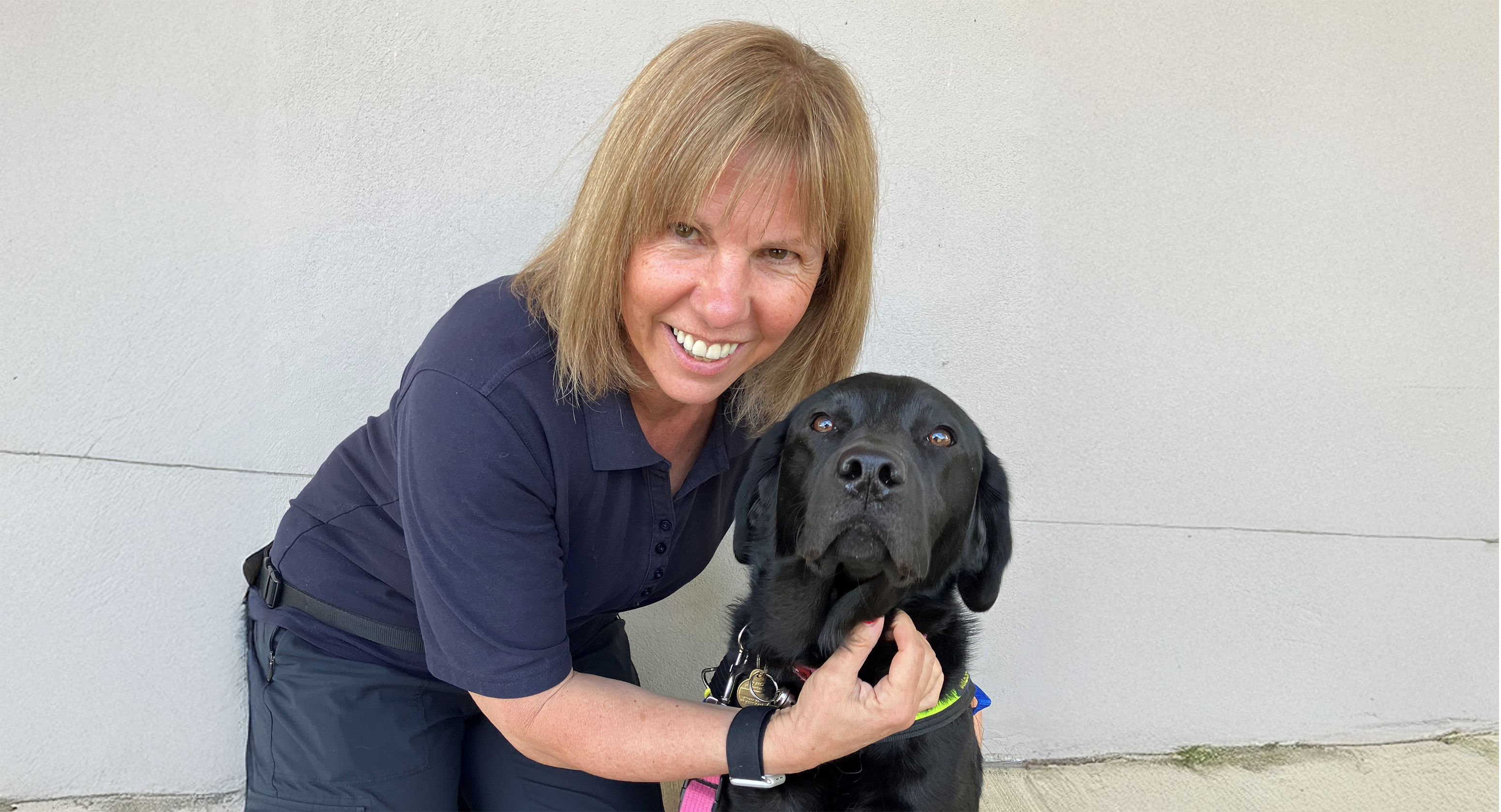 Lucia Corradi is seen kneeling down against a white wall with her Seeing Eye Dog, Luna, next to her. Lucia is in all navy clothes and smiles at the camera, with Luna, a black Labrador, looking directly at the camera as well.