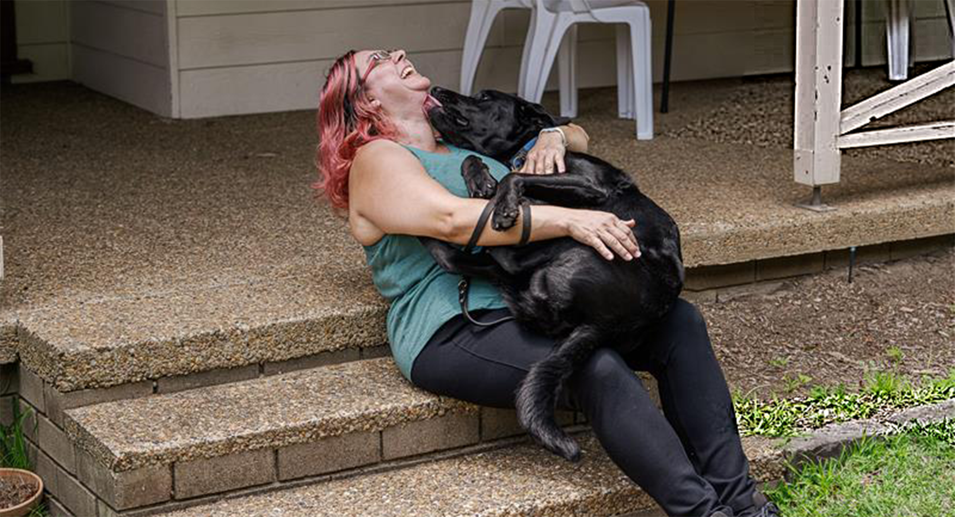 Marilena Osborne is pictured sitting on some stairs and cradling her Seeing Eye Dog, Hamilton. She has her head thrown back in laughter as Hamilton, a black Labrador, lovingly licks her face.