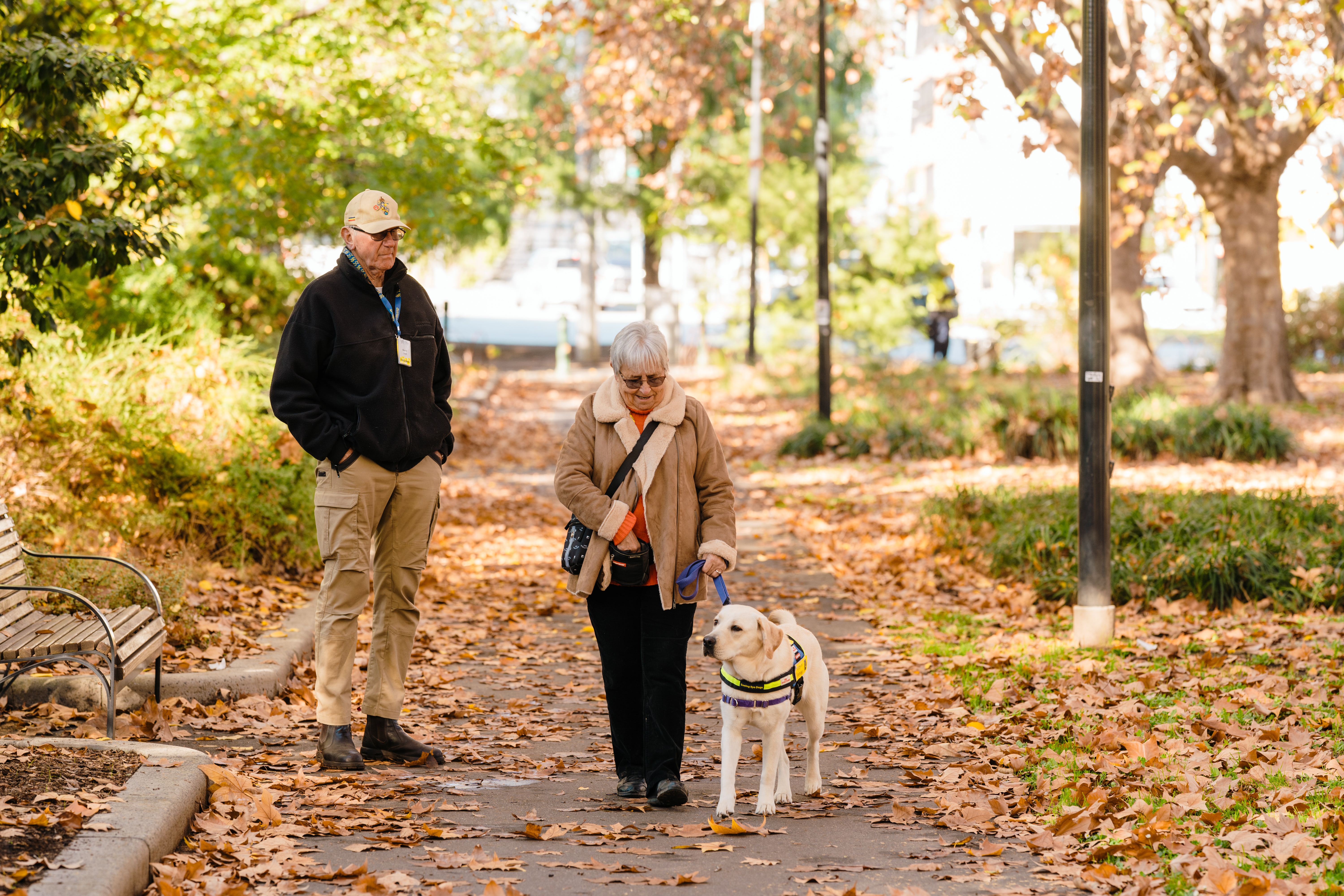 Marilyn leads a yellow Seeing Eye Dogs puppy down a leafy path in a park with her husband standing to her right. 