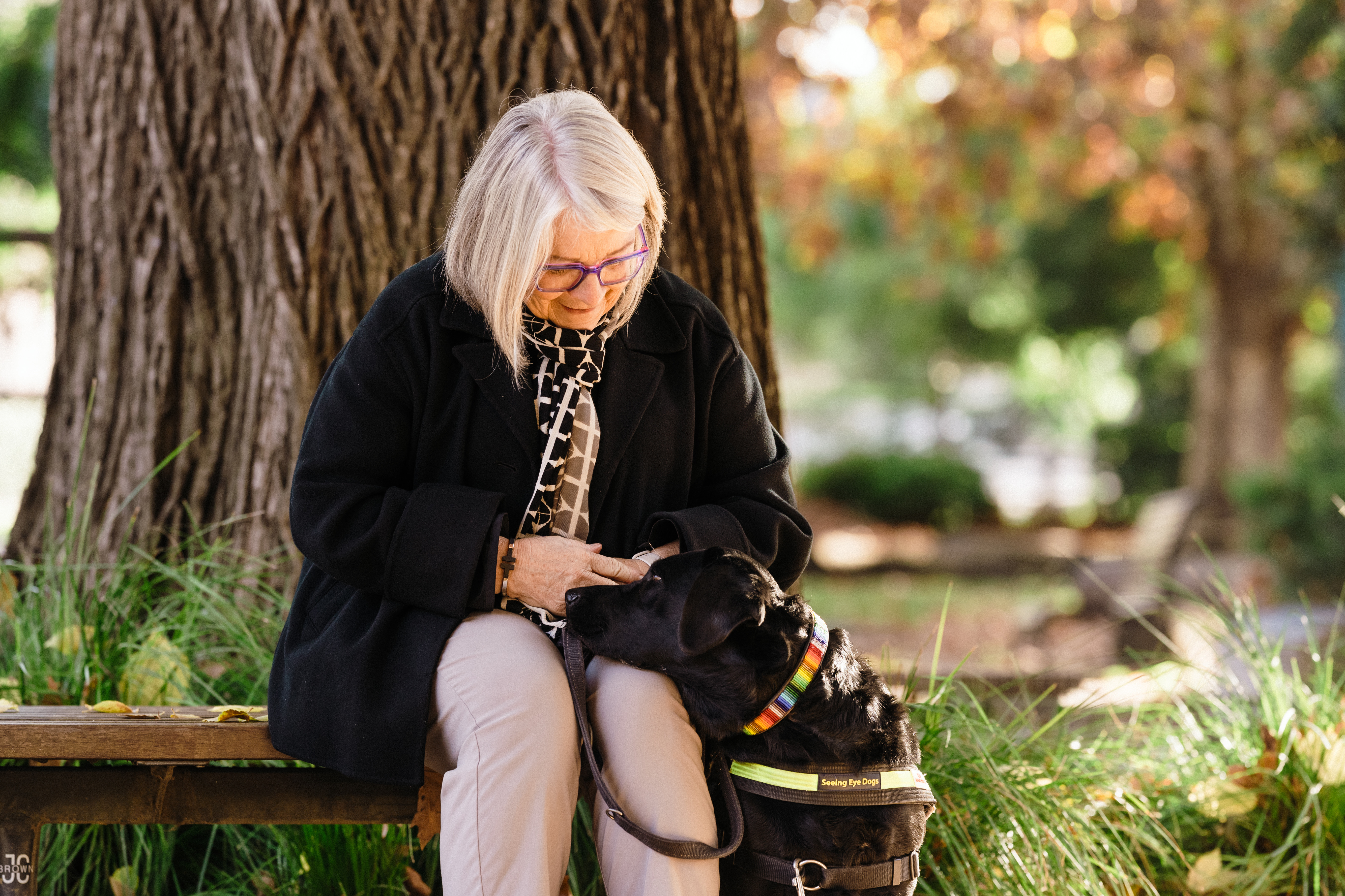 Moira sits on a park bench with black Seeing Eye Dog Natasha sitting next to her, resting her chin on Moira’s knee. 