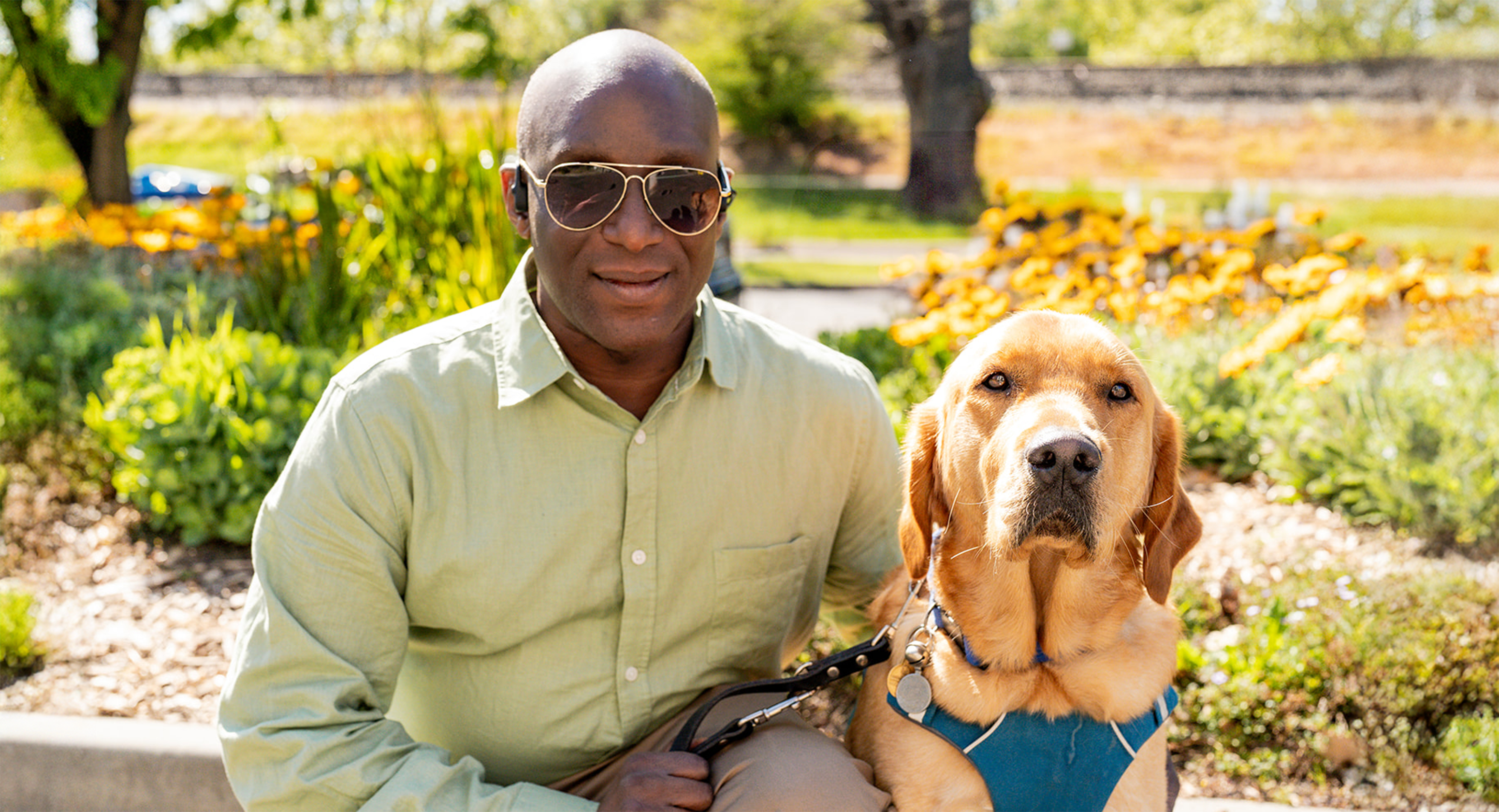 Nemoy Malcolm is kneeling next to his Golden Retriever Seeing Eye Dog, Benji, on a sunny day. He is wearing sunglasses and a light green shirt, while smiling at the camera. Benji sits calmly beside him looking at the camera, wearing a teal harness. They are surrounded by green plants and bright yellow flowers.