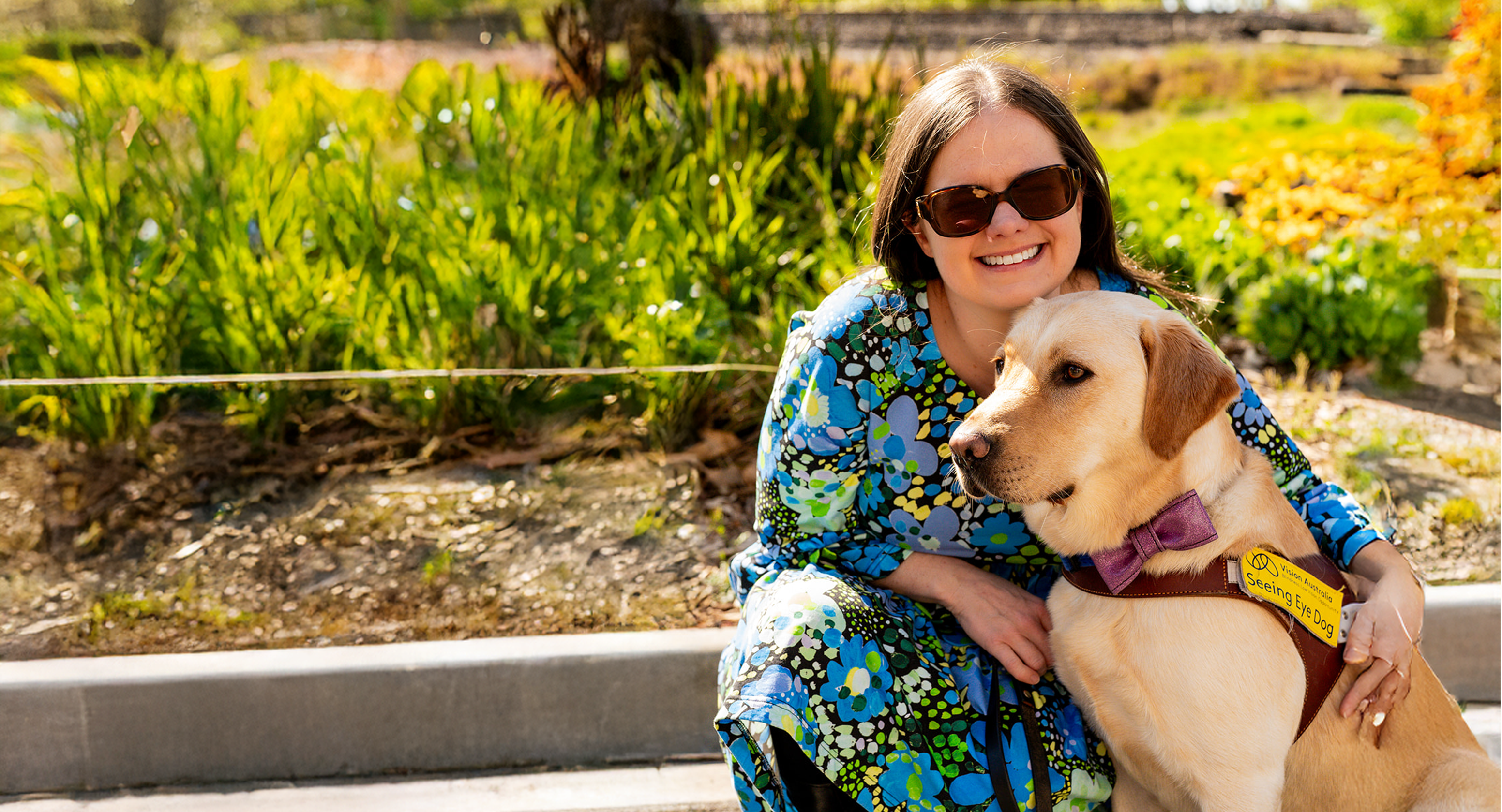 Nicole Tilltoson is kneeling next to her Yellow Labrador Seeing Eye Dog, Trixie, on a sunny day. Nicole is wearing sunglasses and a bright floral patterned blue and green dress, while smiling at the camera. Trixie is wearing a sparkly pink bow tie and is slightly angled away from the camera. They are surrounded by green foliage.