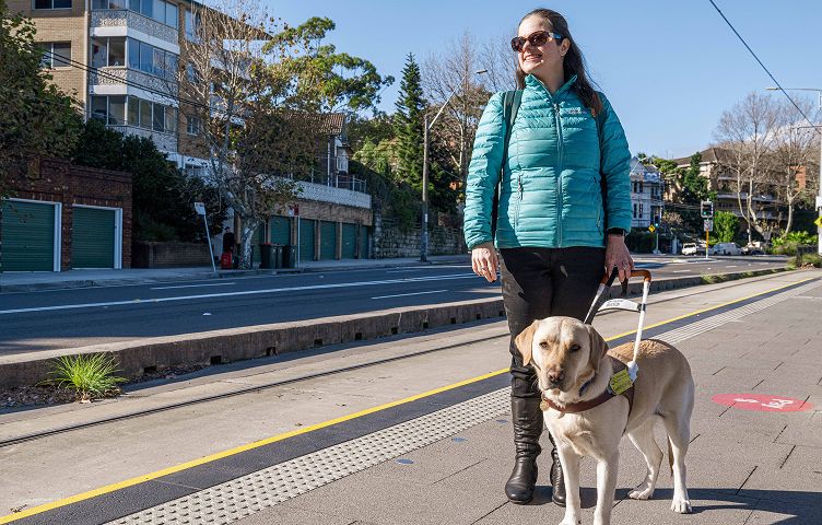 Nicole is standing on a tram platform, she is wearing a puffy jacket and sunglasses, and holding the harness of yellow Seeing Eye Dog Labrador, Trixie.