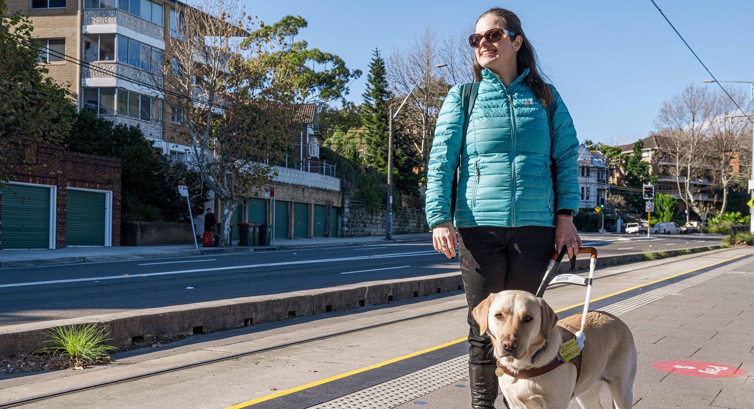 Nicole is standing on a tram platform, she is wearing a puffy jacket and sunglasses, and holding the harness of yellow Seeing Eye Dog Labrador, Trixie.