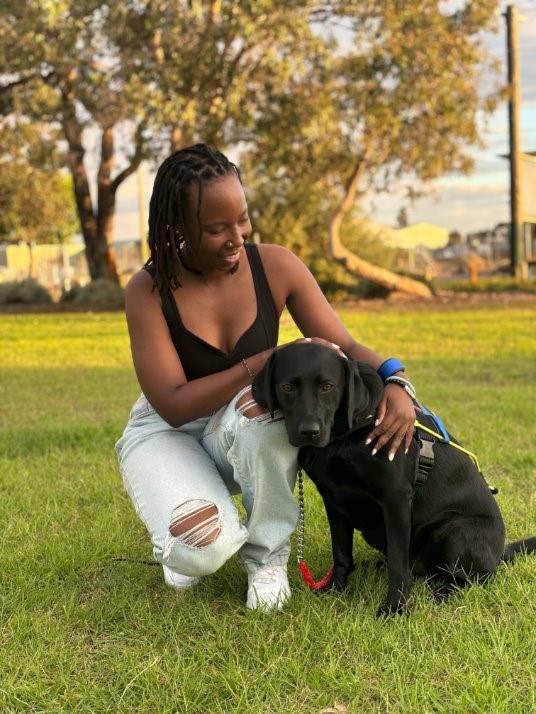 Thayana is kneeling on the ground, with her Seeing Eye Dog Shaylah sitting beside her