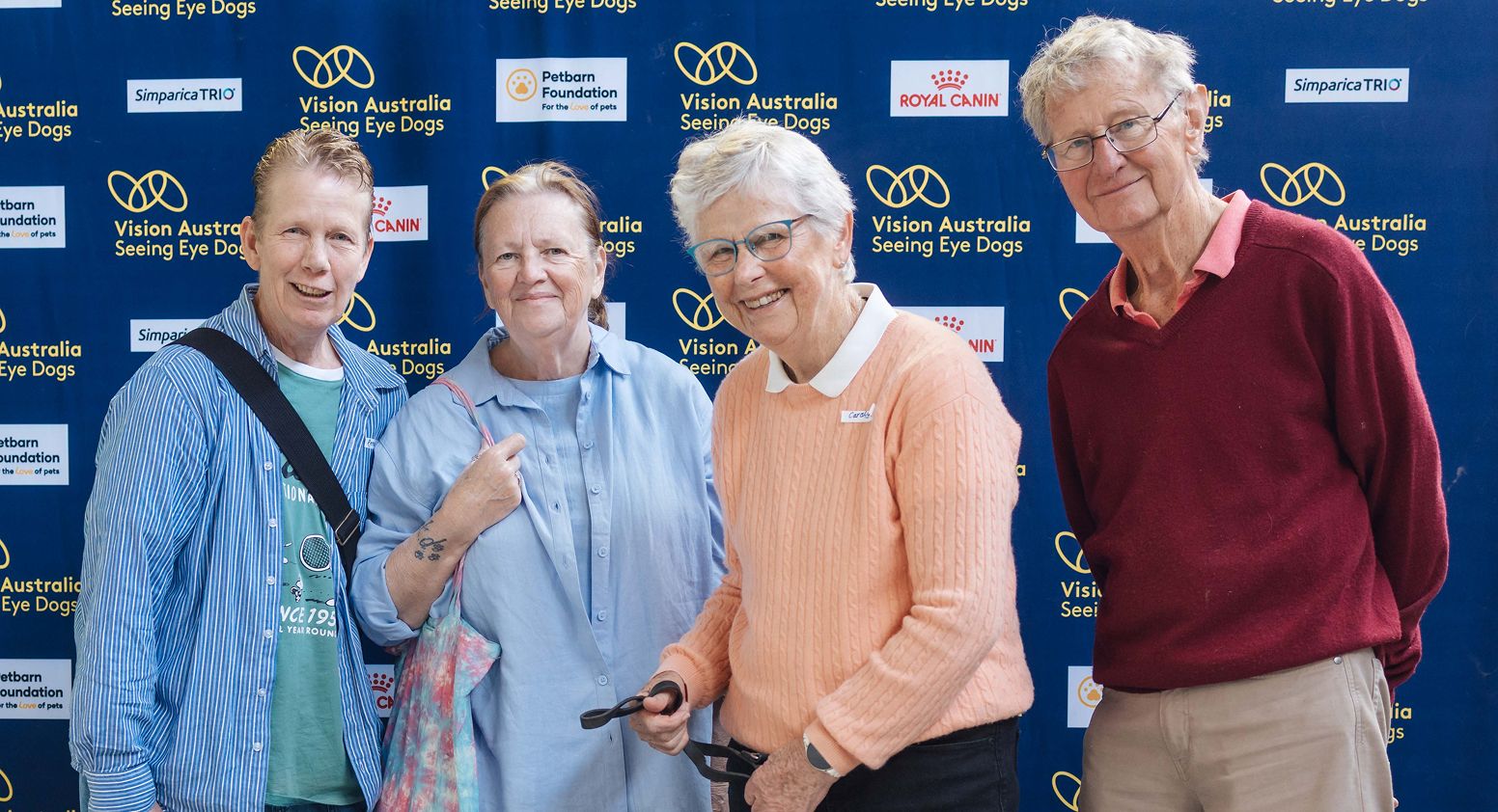 Four smiling puppy sponsors pose together in front of a blue "Vision Australia Seeing Eye Dogs" media backdrop with a yellow Labrador Seeing Eye Dog