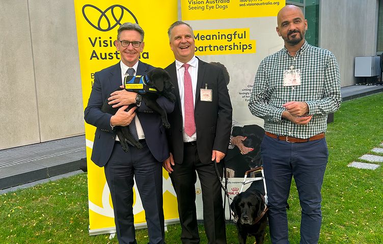 David Williamson, Chris Edwards and Lester Chraim standing and smiling outdoors on a grassy lawn in front of bright yellow Vision Australia Seeing Eye Dogs banners.