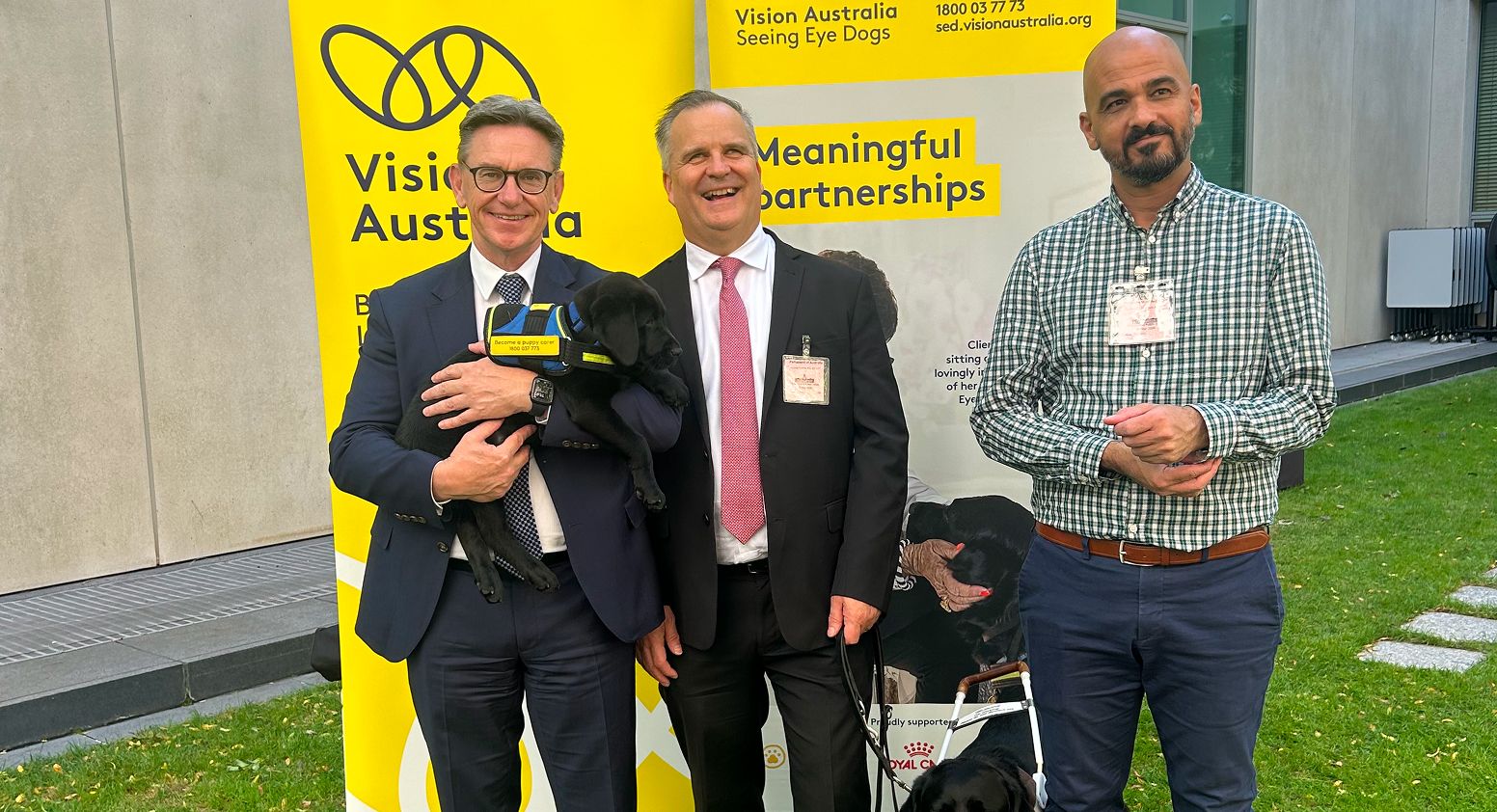 David Williamson, Chris Edwards and Lester Chraim standing and smiling outdoors on a grassy lawn in front of bright yellow Vision Australia Seeing Eye Dogs banners.