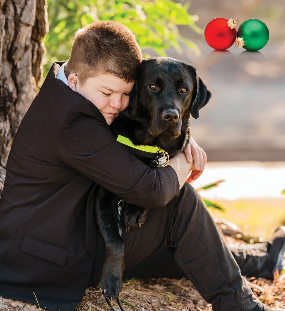 A person in a black coat is sitting on the ground, hugging a black Seeing Eye Dog wearing a yellow harness.