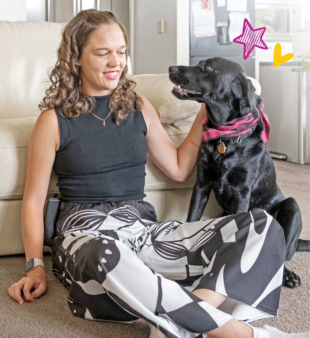 A woman is seated on the ground, in front of a couch and beside her is a black Labrador Seeing Eye Dog