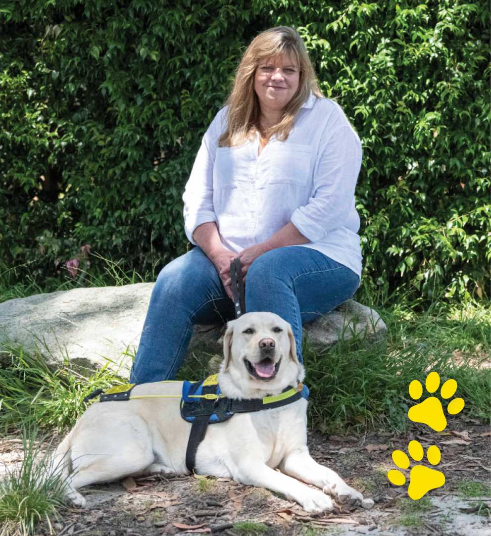 A woman in a white button-up shirt sitting on a rock in a garden. A yellow Seeing Eye Dog is sitting on the ground at her feet.