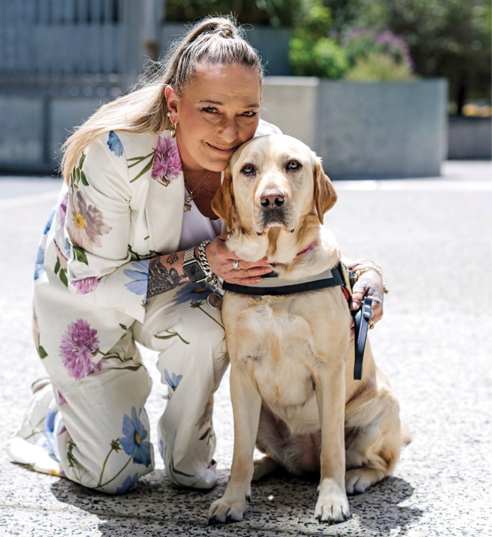 Sally wears a floral suit and is kneeling down, leaning her head affectionately against her Seeing Eye Dog, Joyce.