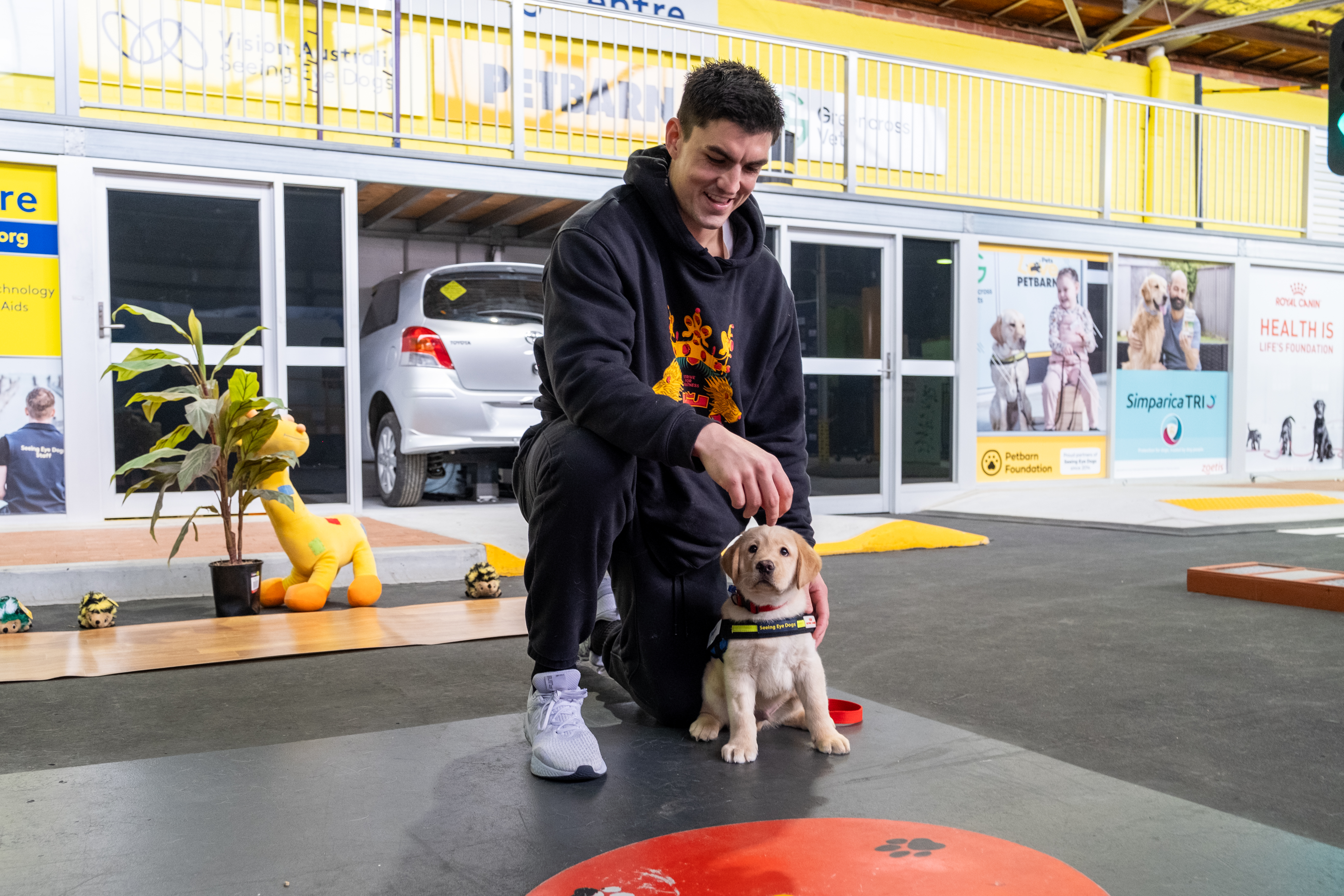 "Brayden Maynard smiling and crouched down next to a yellow Seeing Eye Dogs puppy in jacket at our training facility."