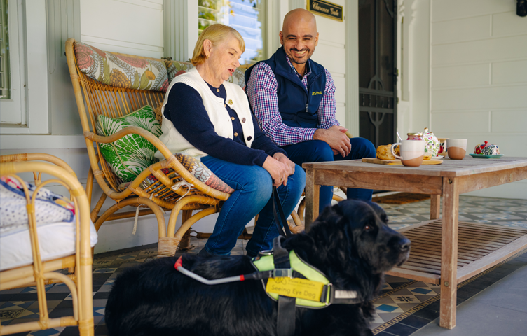 A woman is seated on an patio, next to a Seeing Eye Dog staff member, a black Seeing Eye Dog in harness is laying in on the ground in front.