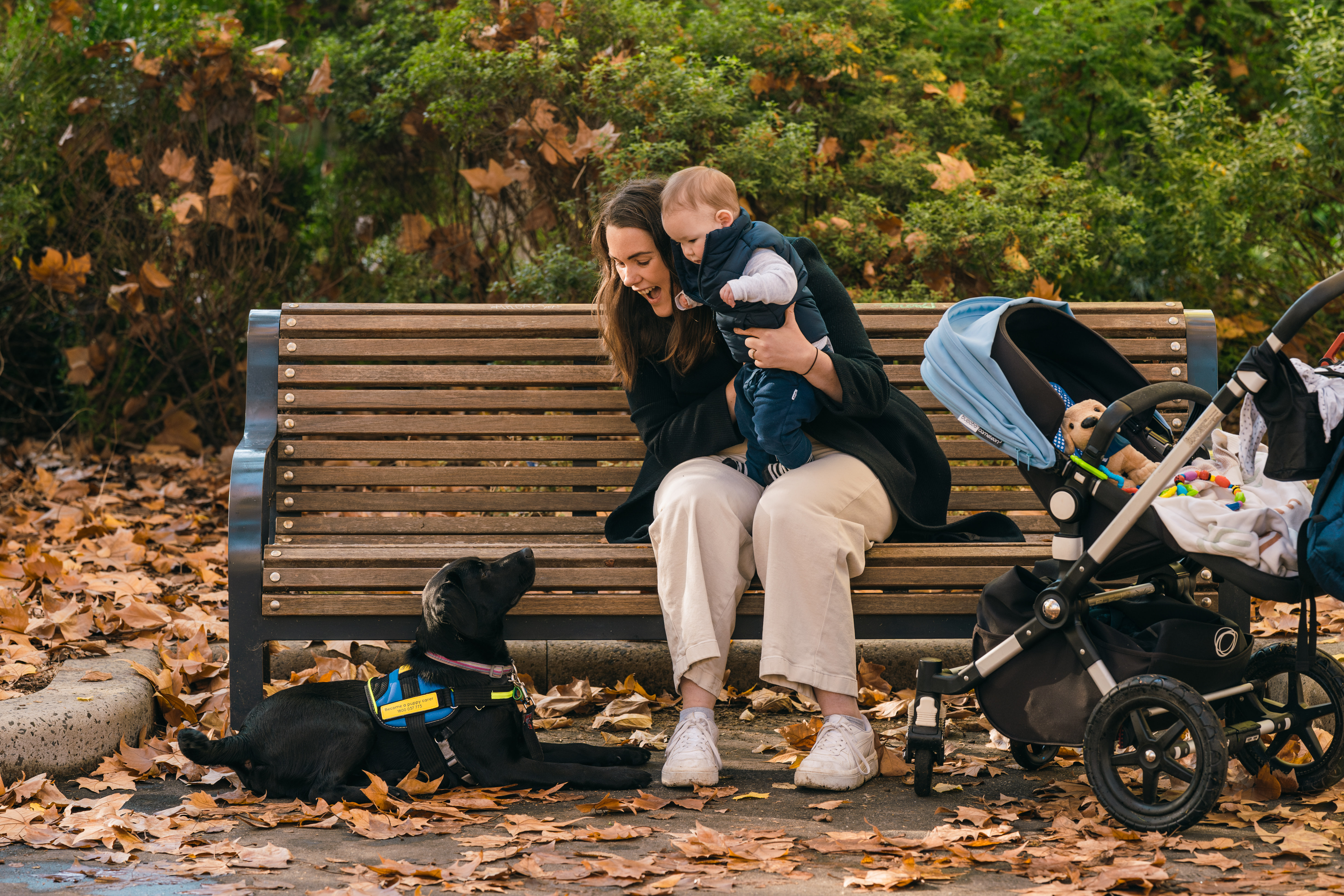 Tamara sits on a wooden park bench holding her baby and smiling down at a black Seeing Eye Dogs puppy, who is lying down next to them and looking up at the baby. 