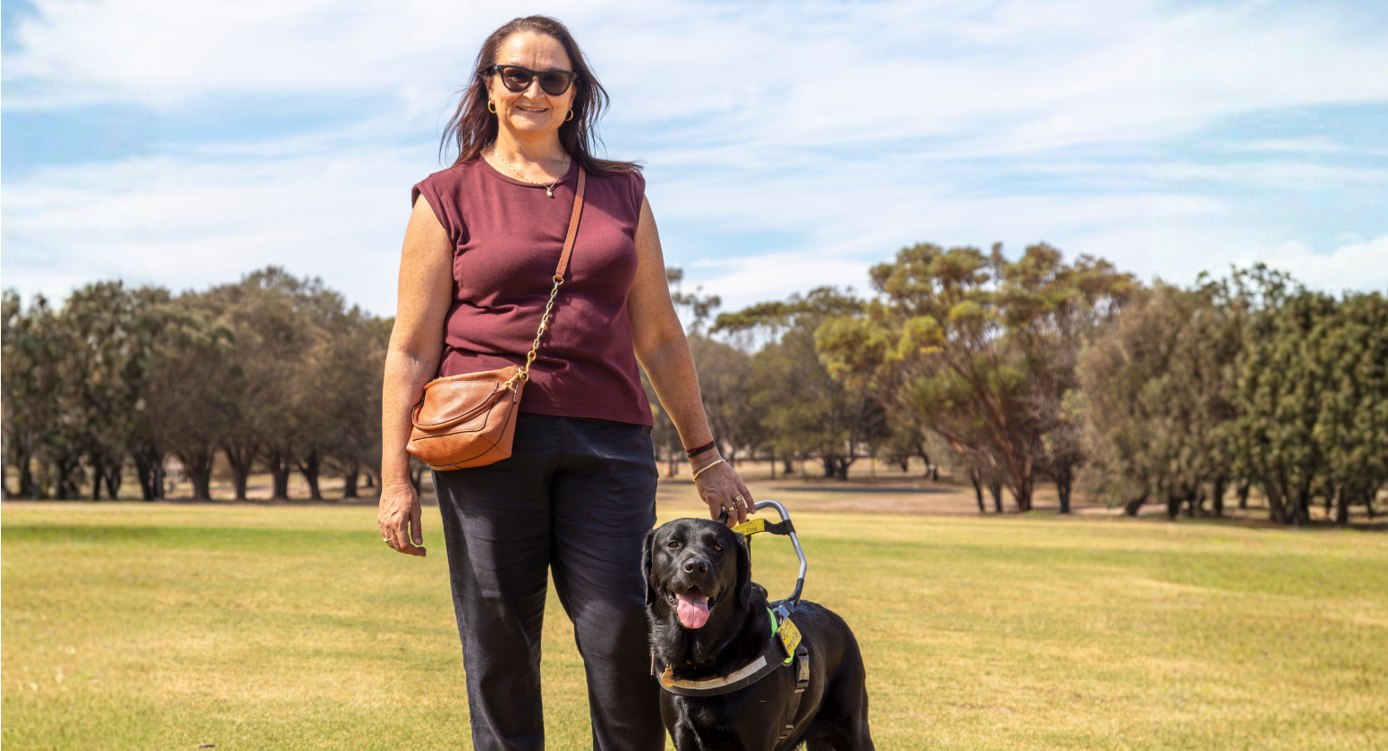 Seeing Eye Dogs client, Tarina, stands next to her black Labrador Seeing Eye Dog, Mika, while smiling at the camera. The two stand in front of a large rural property with trees and grass in the background.