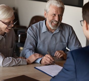 An older man and an older woman are sitting at a desk. They are about to sign a document,
