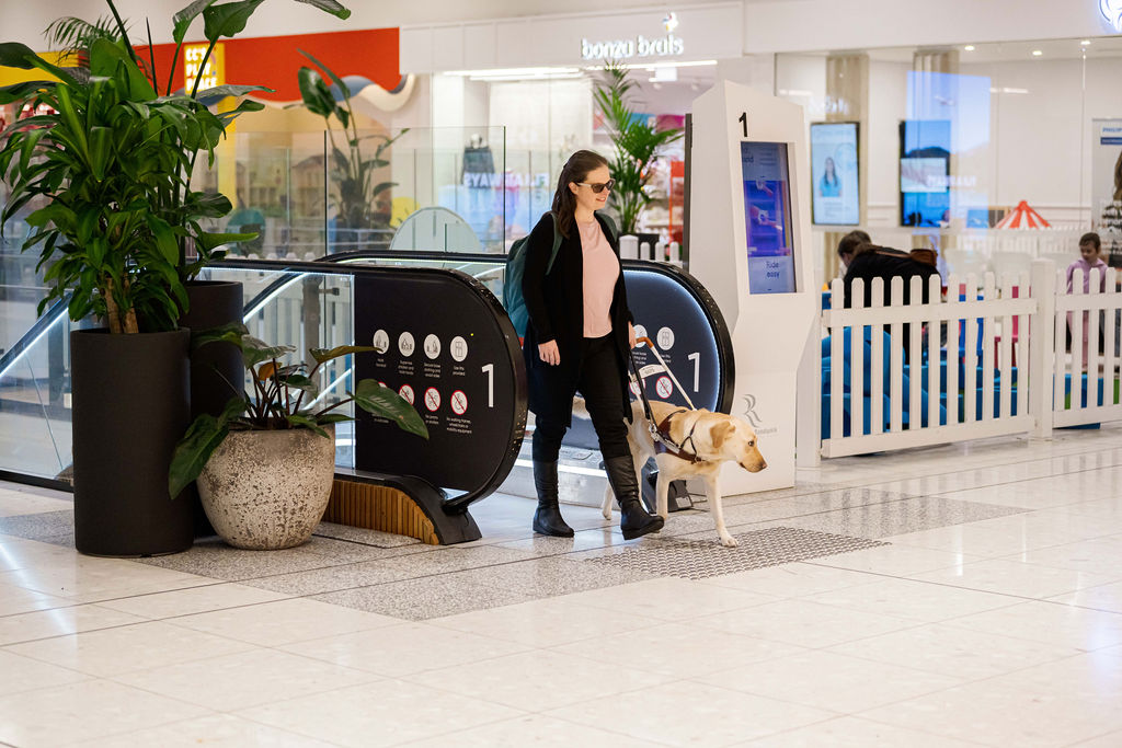 A lady walking off an escalator, accompanied by a Seeing Eye Dog.