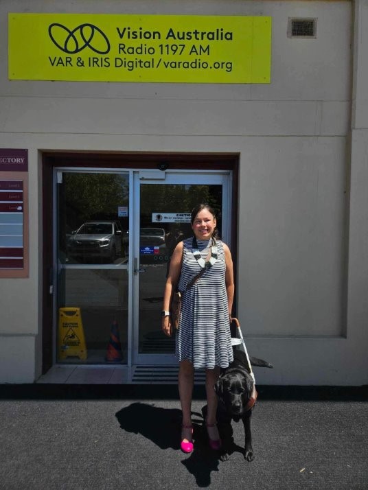 Lizzie is standing outside the Vision Australia Radio offices, with her Seeing Eye Dog Lacey seated beside her. 