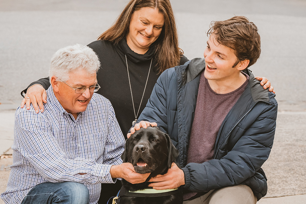 Client Harry sitting with his family and hugging his black Seeing Eye Dog, Bateman