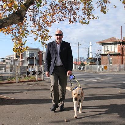 Ron Hooton wearing blacked out glasses, walking along a footpath with a trained Seeing Eye Dog