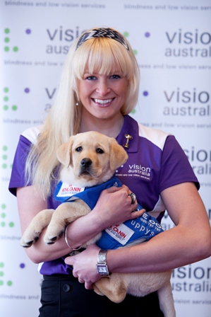 Jess Gallagher smiling and holding a Seeing Eye Dog puppy in front of Vision Australia poster in background.
