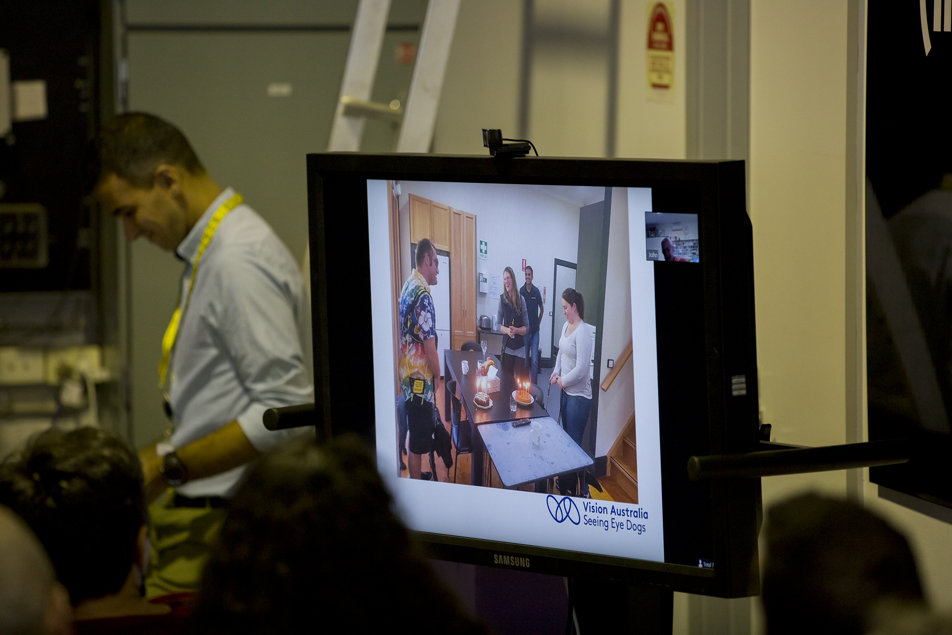 "Graduates and other staff members on a television screen enjoying the celebrations around a table with a cake on it."