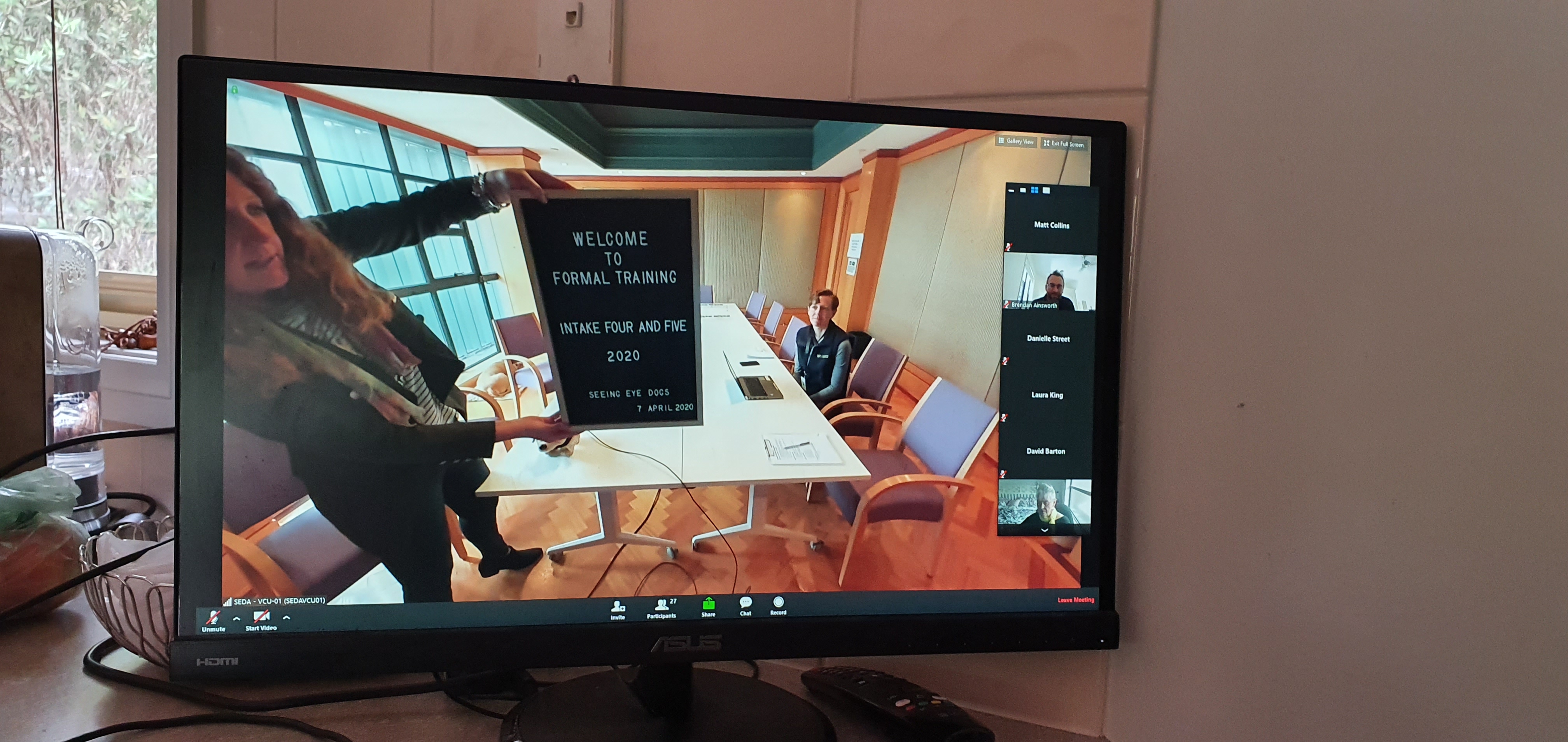 A computer screen joining the videoconference shows Puppy Development Manager Jane holding up a sign reading "Welcome to formal training, Intake Four and Five, 7th April 2020 
