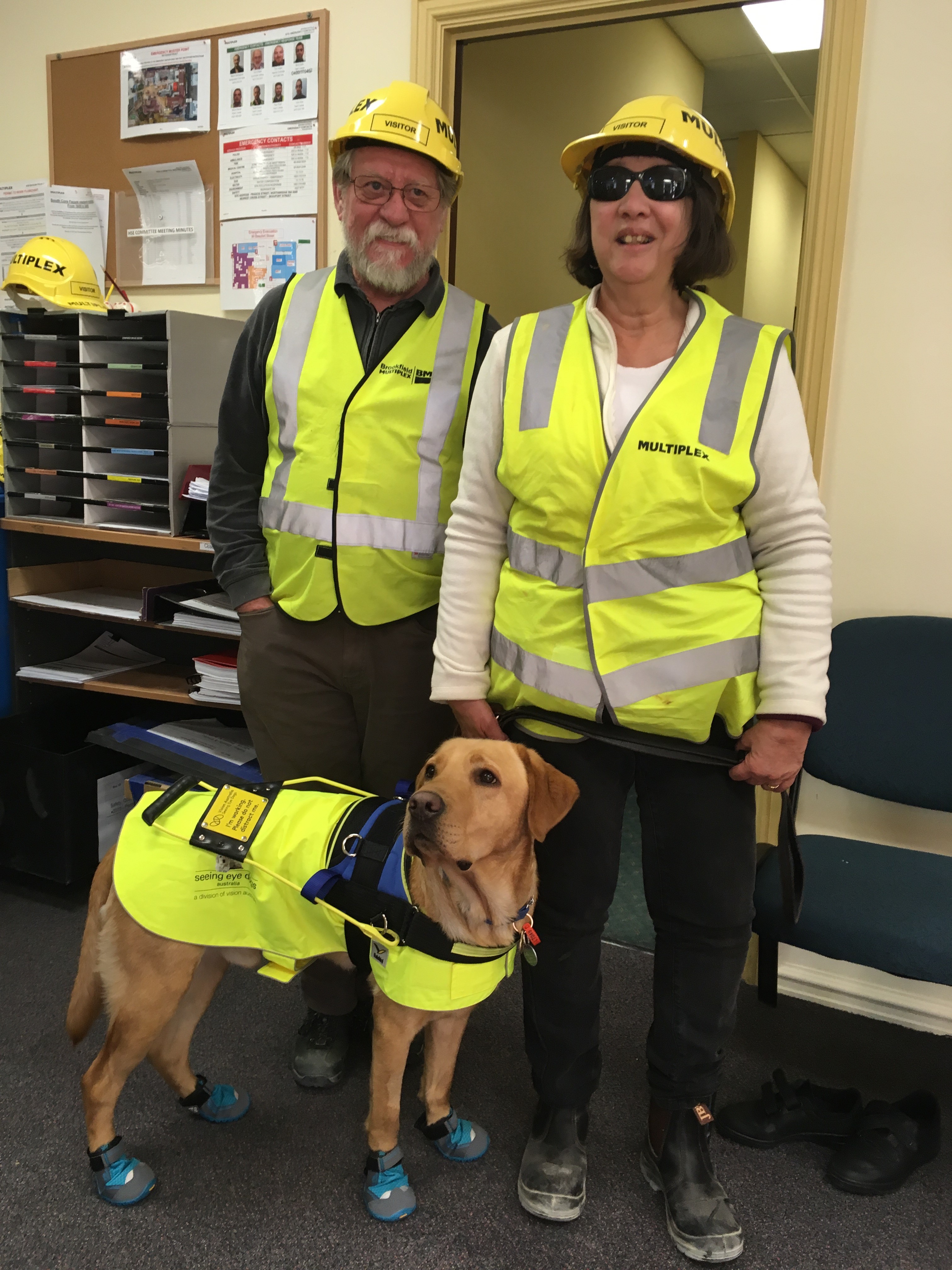 Pamela and her husband stand in high-visibility vests behind Pamela's Seeing Eye Dog Jock, who is also wearing a custom high-visibility vest and protective booties