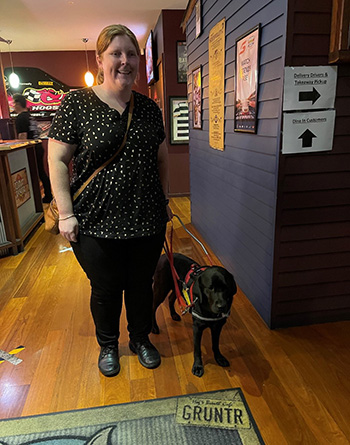 Isabella is smiling at the camera while holding the harness of her black labrador Seeing Eye Dog, Penny